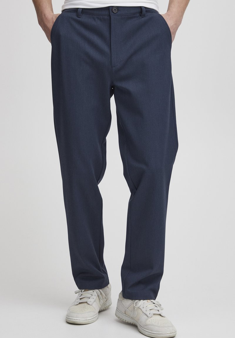 Navy blue tailored trousers with a straight fit, made of a textured fabric. Features a single button closure and side pockets. Worn with white sneakers.