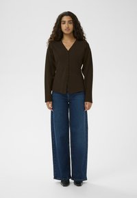 Brauns, garais piedurkņu kardigans ar V-veida kaklu un pogu priekšējo daļu; kombinēts ar plašām, tumši zilām biksēm un melnām kurpēm.