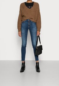 Suéter marrón de punto, blusa negra ajustada, jeans ajustados de tiro alto en azul con desgastes, botines negros y bolso negro.