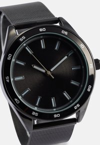 Sort analogur med et struktureret gråt læderrem, minimalistisk urskive, sølvværdi markører og en kontrasterende minutviser mod en mat sort baggrund.