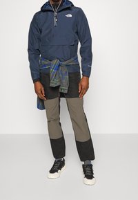 Giacca a vento blu navy con mezza zip, cappuccio e tasca frontale; flanella a quadri sovrapposta in vita; pantaloni bicolore grigio e nero; sneaker nere.