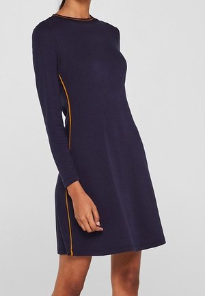 Mujer que lleva un vestido azul marino de manga larga, ajustado y con ribete naranja en los laterales, de pie frente a un fondo gris liso.