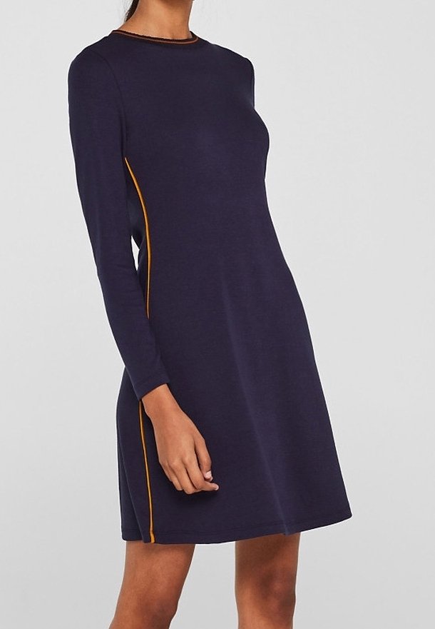 Femme portant une robe bleu marine à manches longues, coupe ajustée avec un passepoil orange sur les côtés, debout devant un fond gris uni.
