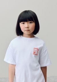 T-shirt en coton blanc à coupe décontractée, avec un petit motif graphique orange sur le côté gauche de la poitrine, représentant des personnages ludiques.