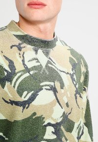 Hombre vistiendo un suéter de cuello redondo con un patrón de camuflaje abstracto en verde, beige y negro, mostrado desde el cuello hasta la parte superior del pecho.