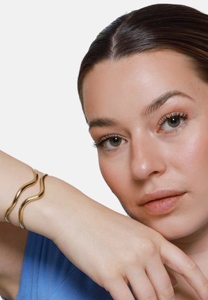 Vrouw met een gladde huid en bruin haar die haar kin op haar opgeheven arm laat rusten, met een gouden golvende armband en een blauwe top.