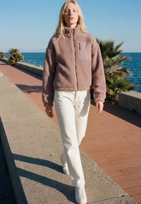 Ung kvinde går på kystpromenaden iført beige fleecejakke, hvide bukser og sneakers under en klar blå himmel.