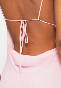 Ružové šaty bez chrbta s tenkými ramienkami a viazaním. Tkanina vyzerá mäkko a hladko, zvýrazňujúc dizajn otvoreného chrbta.