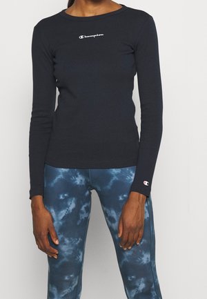 Henkilö, joka pukeutunut mustaan istuvaan pitkähihaiseen Champion-paitaan ja sinisiin kirjaviin leggingsseihin, seisoo yksinkertaista harmaata taustaa vasten.