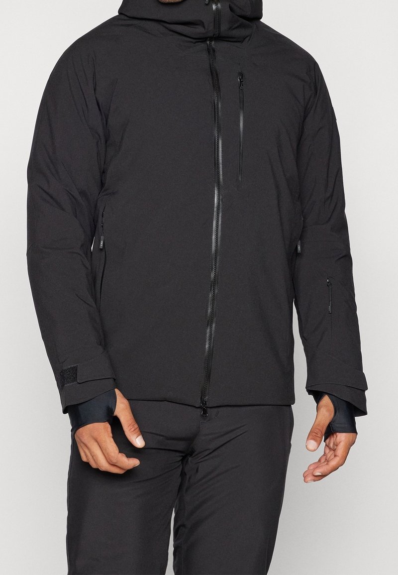 Muž v černé mikině s kapucí a odpovídajících kalhotách se zipovými kapsami a otvorů pro palce, stojící na jednobarevném pozadí.