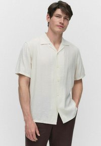 Chemise à manches courtes, col montant en crème clair, fabriquée en tissu texturé. Présente un devant à boutons et une coupe décontractée, assortie à un pantalon marron foncé.