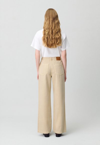 Pantalones anchos beige con una tela texturizada, dos bolsillos traseros y un parche de cuero marrón en la cintura. Combinados con una camiseta blanca.