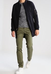 Giacca bomber blu scuro sopra un maglione lavorato a maglia grigio, abbinata a pantaloni verde oliva e sneakers bianche. La giacca presenta una tasca con zip.