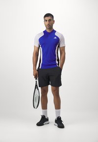 Mannen sportoutfit met een blauwe T-shirt met korte mouwen met witte en zwarte accenten, gecombineerd met zwarte shorts en zwarte sneakers.