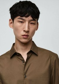 Camicia button-up marrone con colletto, vestibilità slim, texture liscia e bottoni scuri, indossata da un modello con capelli corti e texturizzati.