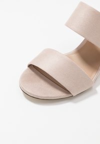 Sandale en daim nude avec deux larges sangles, semelle plate et texture lisse. Design minimaliste sans matériel ou motifs visibles.