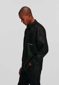 Cămașă din denim negru cu un buzunar și broderie subtilă a logo-ului. Mâneci lungi și guler, având o croială lejeră și o textură moale.