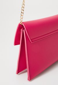 Světle růžová kožená kabelka s překlopeným víkem a krátkým zlatým řetízkovým popruhem, vyfotografovaná na bílém pozadí.