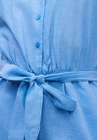 Φόρεμα ανοιχτού μπλε με υφασμάτινη ζώνη δεμένη σε φιόγκο στη μέση και μικρά μπλε κουμπιά στο μπροστινό μέρος.