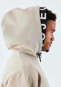 Sweat-shirt à capuche beige avec une doublure intérieure noire imprimée. Il dispose d'un système de fermeture avec cordons et d'un tissu texturé. La vue de profil met en avant les détails de la capuche.