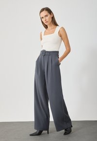 Bredde ben, grå bukser med belte i livet, detaljer med legg, og glatt tekstur, kombinert med en tettsittende hvit tanktop og sorte hæler.