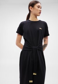 Μαύρο βαμβακερό φόρεμα με κοντά μανίκια, διαθέτει λεπτομέρεια δεμένη στη μέση και χρυσαφί μεταλλικά στοιχεία. Λείο ύφασμα, άνετη εφαρμογή.