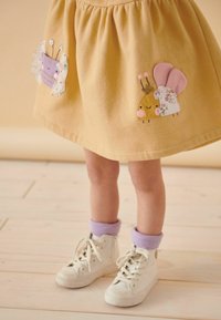 Enfant portant une jupe jaune avec une broderie de insecte souriant, des baskets blanches montantes et des chaussettes violettes sur un sol en bois clair.