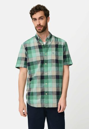 Mand med skæg iført ternet skjorte med korte ærmer i grøn, blå og beige samt mørke bukser, stående mod en hvid baggrund.