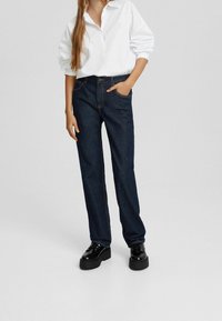 Vaqueros de mezclilla azul oscuro con un diseño de pierna recta, con costuras de contraste y bolsillos frontales, combinados con una camisa blanca de botones extragrande.