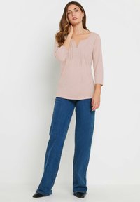 Helles rosa Langarmoberteil mit V-Ausschnitt und gefaltetem Design, kombiniert mit weit geschnittenen blauen Denim-Jeans und schwarzen Schuhen.