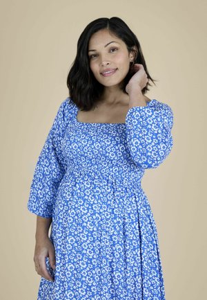 Glunderende vrouw met schouderlang donker haar die een blauwe bloemenjurk met lange mouwen draagt, staand tegen een beige achtergrond, haar haar aanrakend.