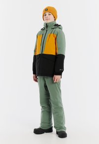 Skijack in groen, zwart en oranje; heeft een ritssluiting aan de voorkant, een capuchon en verstelbare manchetten. Gemaakt met bijpassende groene broek en zwarte laarzen.