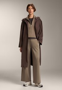Hnedý nepremokavý dlhý kabát cez rebrovaný béžový top a široké nohavice. Topánky sú biele s čiernymi akcentmi. Jednoduchý, strihaný dizajn.