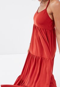 Robe longue couleur rouille avec un décolleté en V, des bretelles tressées ajustables et une jupe à volants à étages, conçue dans un tissu doux et léger.