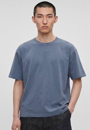 Jeune homme aux cheveux noirs courts portant un t-shirt bleu uni à manches courtes et un pantalon sombre, debout devant un fond gris clair.