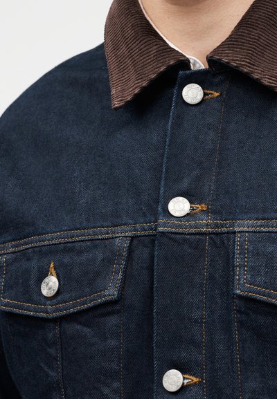 Κοντινή λήψη σκούρας μπλε denim μπουφάν με καφέ γιακά από κορντό και ασημένια κουμπιά, με πορτοκαλί ραφές.