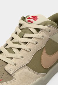 Κοντινή λήψη μπεζ και ελιάς αθλητικού παπουτσιού Nike SB Force 58 με κρεμ κορδόνια και λογότυπο Nike σε ροδακινί χρώμα στο πλάι.