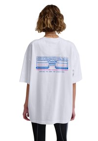 White oversized t-shirt featuring a graphic print on the back, depicting a stadium with blue outlines and text, “SERVING THE GAME FOR GENERATIONS.”