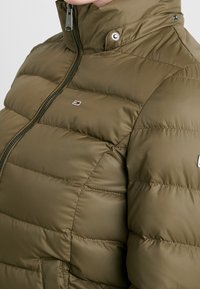 Chaqueta acolchada de color verde oliva con paneles horizontales cosidos, un cuello alto y una cremallera. Presenta un pequeño logo en el pecho y botones a presión.