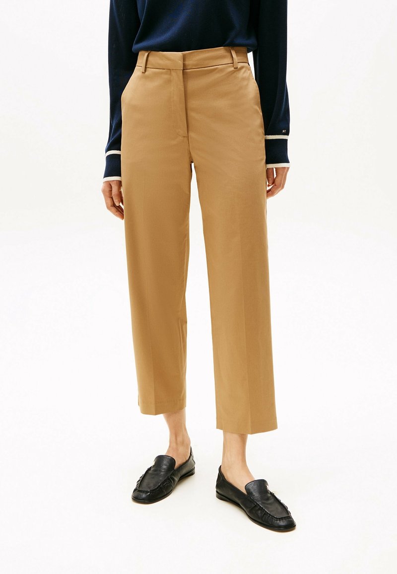 Personne portant un pantalon beige raccourci à jambes droites, des mocassins noirs et un haut à manches longues foncé avec des poignets blancs.
