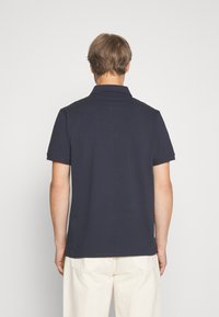 Homme vu de dos portant un polo bleu marine foncé à manches courtes et un pantalon crème clair sur un fond uni.
