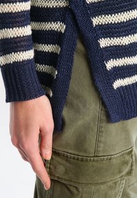 Navyblå och krämrandig stickad cardigan med ribbad manschett, sidosprund och textilstruktur, i kombination med olivgröna cargobyxor.