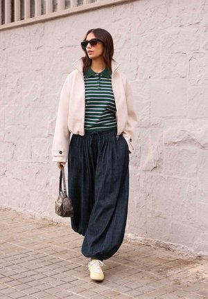 Femeie purtând ochelari de soare, jachetă crem, polo cu dungi verzi, pantaloni largi închiși la culoare, adidași și o geantă metalică mică, mergând pe lângă un zid de piatră.