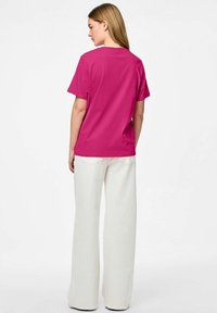 Femme debout, tournée de dos, portant un chemisier rose vif à manches courtes et un pantalon blanc taille haute à jambes larges sur fond blanc.