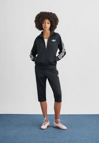 Svart treningsdress med hvite tre-stripe detaljer, inkludert en jakke med glidelås og knelange bukser. Brukt med lyserosa ballettsko.