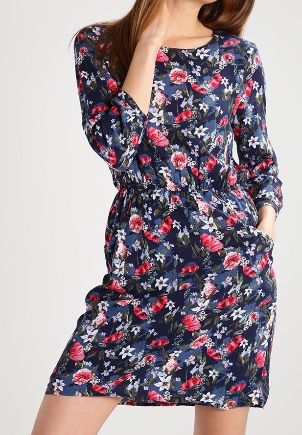 Femme portant une robe fleurie bleu marine avec des fleurs roses, blanches et rouges, manches trois quarts, taille élastique, main dans la poche.