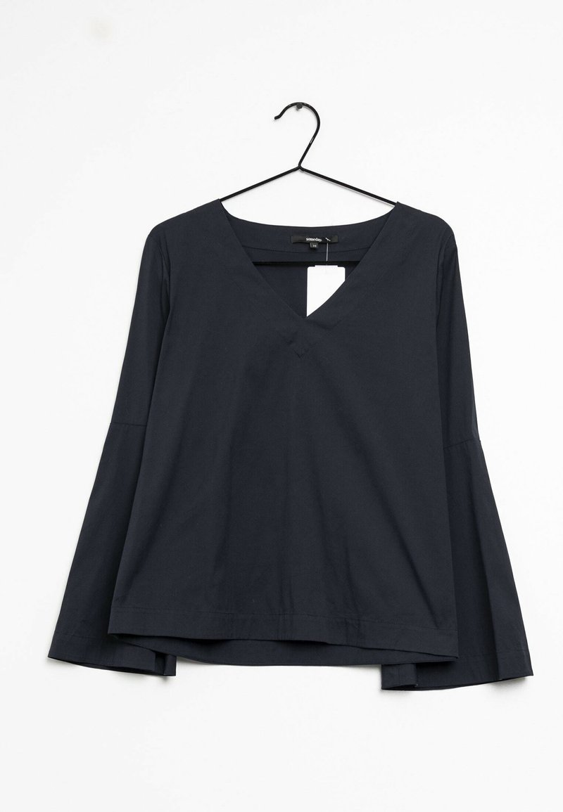 Blouse noire à manches longues avec col en V suspendue sur un cintre noir sur un fond blanc avec une étiquette de prix blanche visible.