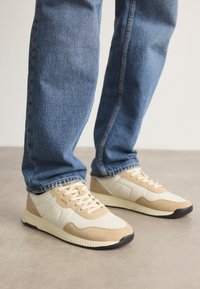Baskets beiges et crème en matériaux suédé et en maille, avec une semelle texturée et des accents ton sur ton, associées à un jean en denim bleu.