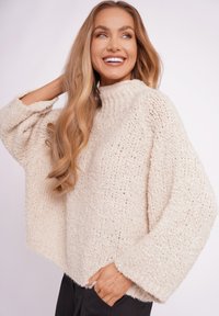 Jasno beżowy sweter dziany z wysokim dekoltem, szerokimi rękawami i luźnym krojem, wyróżniający się fakturowanym wzorem i miękkim wykończeniem.