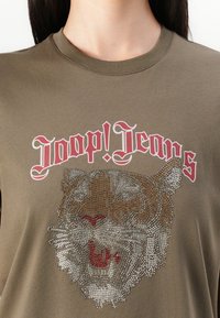 Olivenfarget bomulls T-skjorte med et trykk av en tiger prydet med rhinestones og "Joop! Jeans" tekst i rødt. Rund hals og korte ermer.
